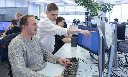 Ein Mann sitzt in einem Großraumbüro an einem PC; neben ihm steht ein junger Mann, der auf einen Bildschirm zeigt.